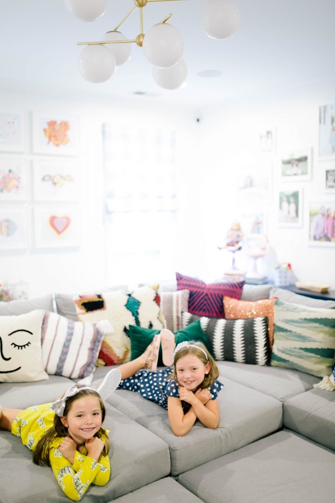 Fashionistas in Training + Nordstrom Kids Clothing by popular Nashville life and style blog, Hello Happiness: image of two girls laying on their stomachs on a grey sectional couch and wearing a Nordstrom Tea Collection Ruffle Dress, Native Shoes Jefferson Bling Glitter Slip-On Vegan Sneaker, Tucker + Tate Wide Leg Romper, Maniere Metallic Headband, and Mel by Melissa Cosmic Sandal. 