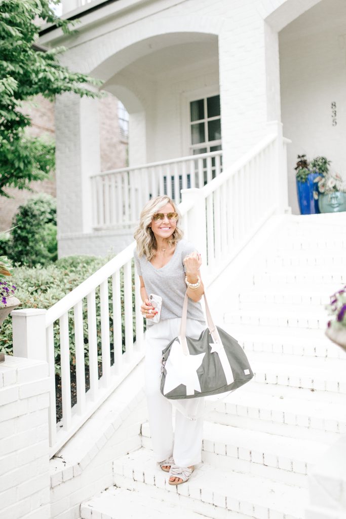 We Dress America + Walmart Travel Style for a Successful Getaway by top US fashion blog, Hello Happiness: image of woman standing outside on white brick steps wearing a gray v-neck t-shirt, sunglasses, white pants, stripe slide sandals, and holding a large gray duffle bag with a white star print and white straps.
