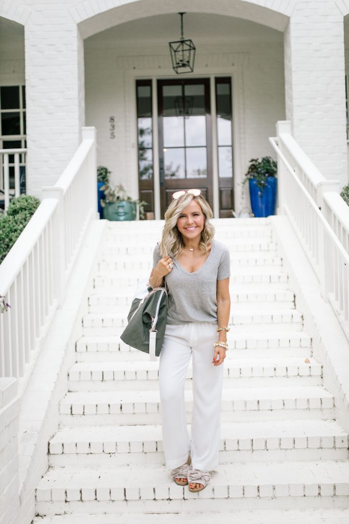 We Dress America + Walmart Travel Style for a Successful Getaway by top US fashion blog, Hello Happiness: image of woman standing outside on white brick steps wearing a gray v-neck t-shirt, sunglasses, white pants, stripe slide sandals, and holding a large gray duffle bag with a white star print and white straps.