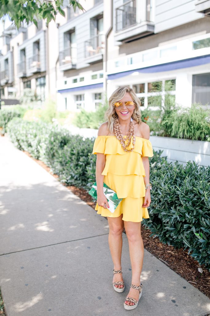 Those Hot Summer Nights... A New Collection from Gibson! by popular Nashville fashion blog Hello Happiness: image of a woman standing outside on a sidewalk next to a white building wearing a yellow Gibson Hot Summer Nights Natalie Off The Shoulder Ruffle Dress, Hearne Dry Goods Co. Tahiti Beach necklace, Steve Madden Bree Leopard wedge sandals, and holding a H. Howorth Art Emerald Green Clutch.
