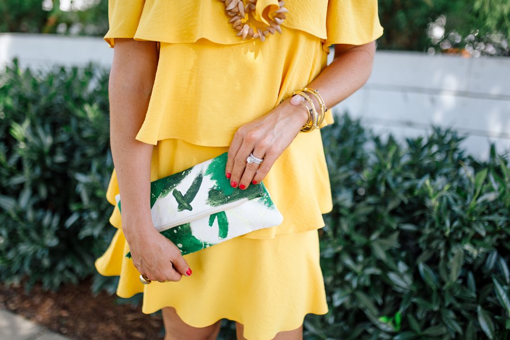 Those Hot Summer Nights... A New Collection from Gibson! by popular Nashville fashion blog Hello Happiness: image of a woman standing outside on a sidewalk next to a white building wearing a yellow Gibson Hot Summer Nights Natalie Off The Shoulder Ruffle Dress, Hearne Dry Goods Co. Tahiti Beach necklace, Steve Madden Bree Leopard wedge sandals, and holding a H. Howorth Art Emerald Green Clutch.