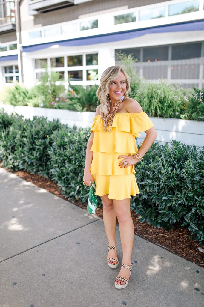 Those Hot Summer Nights... A New Collection from Gibson! by popular Nashville fashion blog Hello Happiness: image of a woman standing outside on a sidewalk next to a white building wearing a yellow Gibson Hot Summer Nights Natalie Off The Shoulder Ruffle Dress, Hearne Dry Goods Co. Tahiti Beach necklace, Steve Madden Bree Leopard wedge sandals, and holding a H. Howorth Art Emerald Green Clutch.