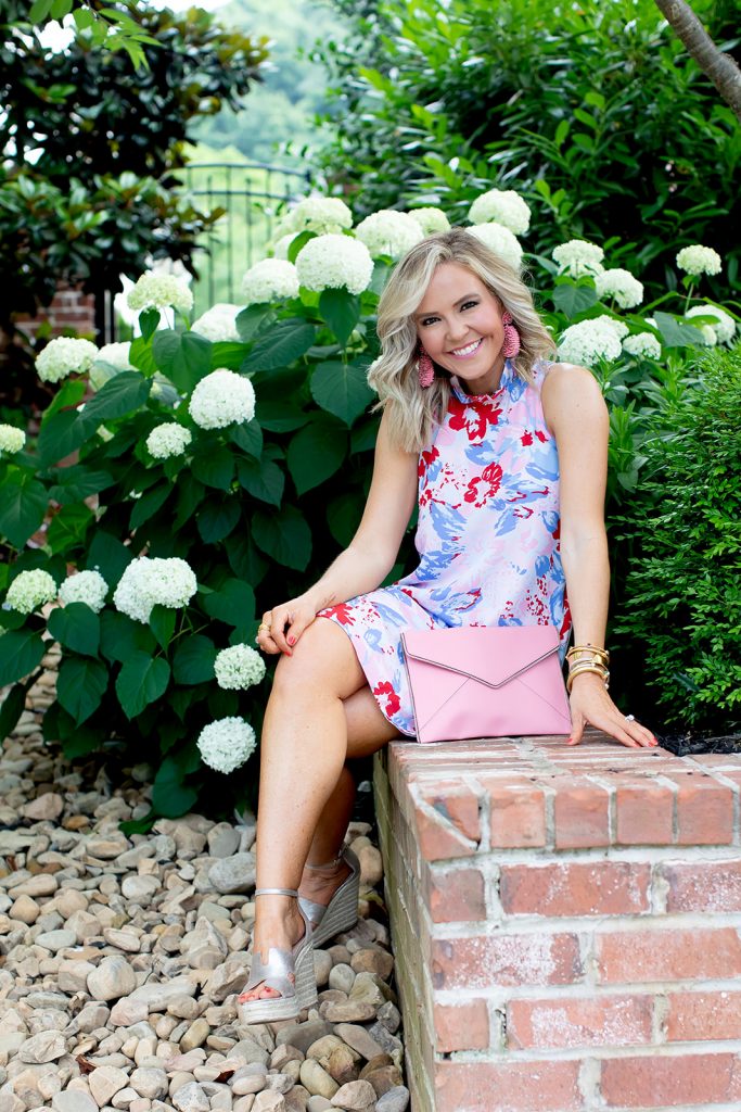 Gibson x Hi Sugarplum Summer of Color Collection by top us fashion blog, Hello Happiness: image of smiling woman sitting outside by a white flowering bush and wearing the Sardinia Ruffle Neck Date Dress in Vista Cherry, gold wedges, beaded earrings, and holding the pink Leo clutch.