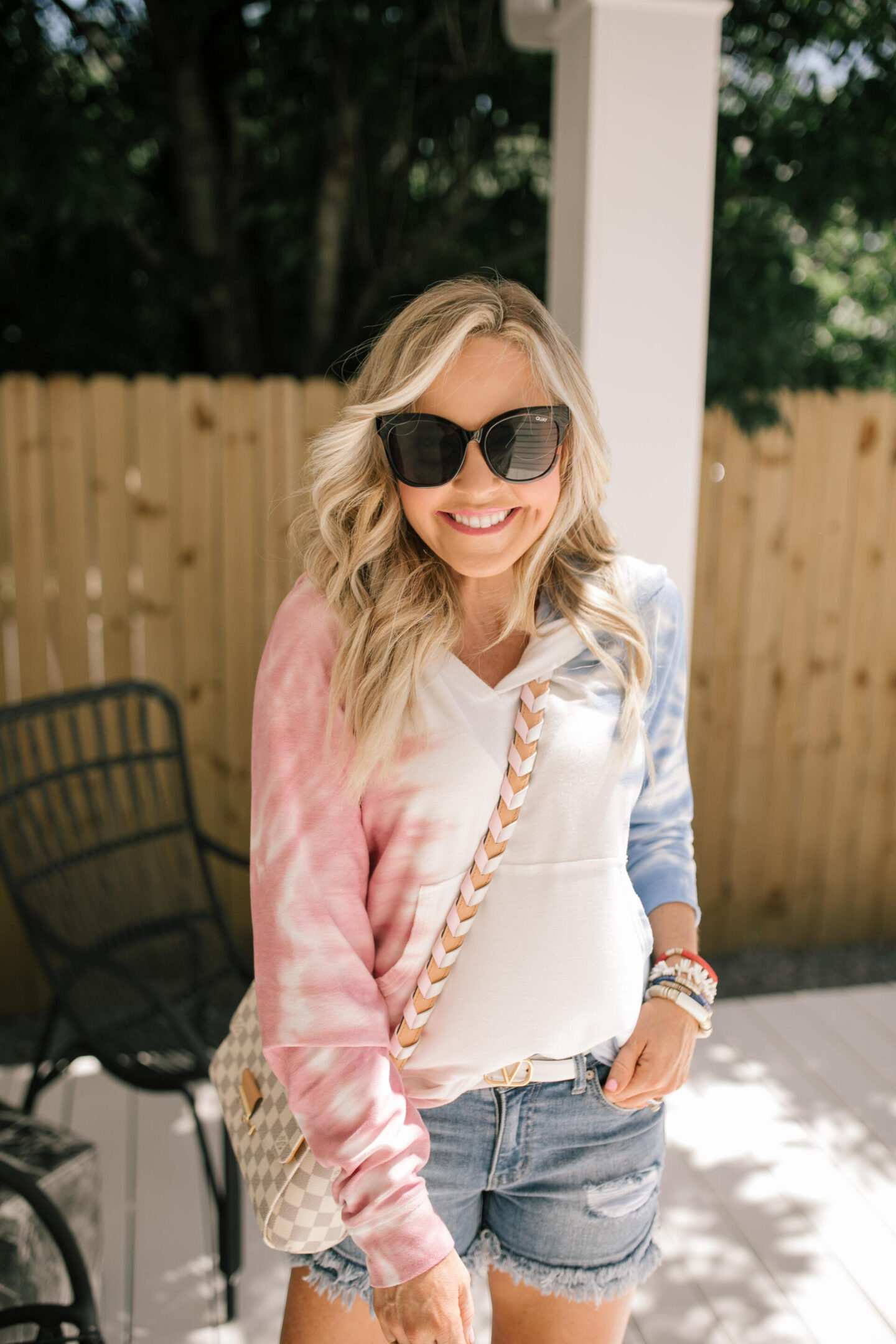 4th of July Outfits by popular Nashville fashion blog, Hello Happiness: image of a woman wearing a red, white, and blue tie dye hoodie, distress hem denim shorts, white Louis Vuitton belt, silver and white sneakers, black frame sunglasses, and holding a grey and white checkered handbag. 