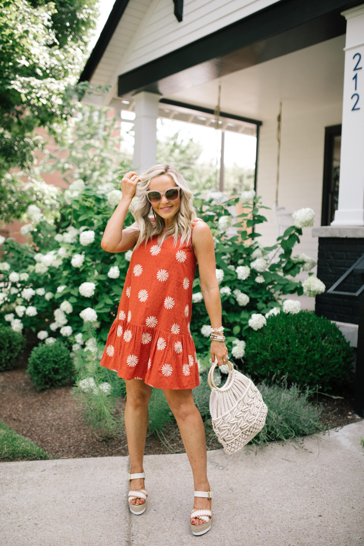 Nashville Hot Spots by popular Nashville travel blog, Hello Happiness: image of Natasha Stoneking wearing a orange sunglower dress, white strap espadrilles, and holding a macrame hand bag while standing next to a white flowering bush. 