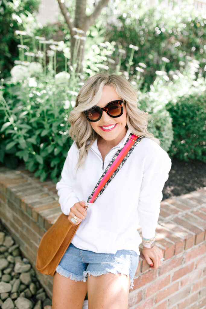 Social Threads by popular Nashville fashion blog, Hello Happiness: image of Natasha Stoneking wearing a Social Threads Ocean Drive Half Zip Sweatshirt, Social Threads Free People CRVY High Rise Cut Offs, Steve Madden TORIN WHITE WITH STUDS sneakers, and carrying a Social Threads Vegan Messenger Bag with a Social Threads Hello Happiness Blogger Collaboration Bag Strap.