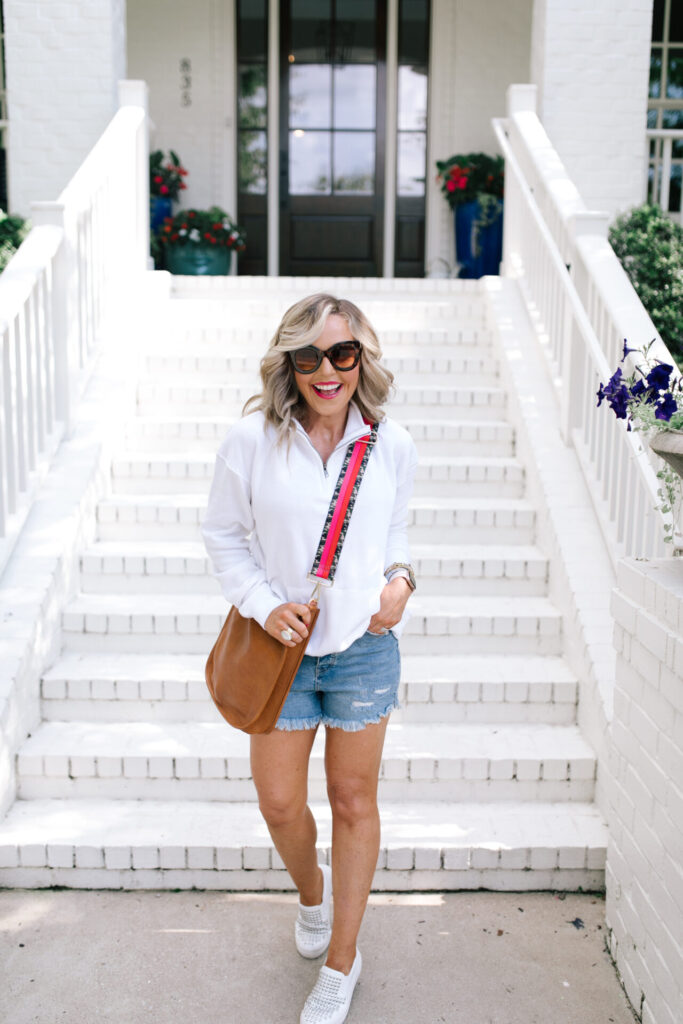 Social Threads by popular Nashville fashion blog, Hello Happiness: image of Natasha Stoneking wearing a Social Threads Ocean Drive Half Zip Sweatshirt, Social Threads Free People CRVY High Rise Cut Offs, Steve Madden TORIN WHITE WITH STUDS sneakers, and carrying a Social Threads Vegan Messenger Bag with a Social Threads Hello Happiness Blogger Collaboration Bag Strap.