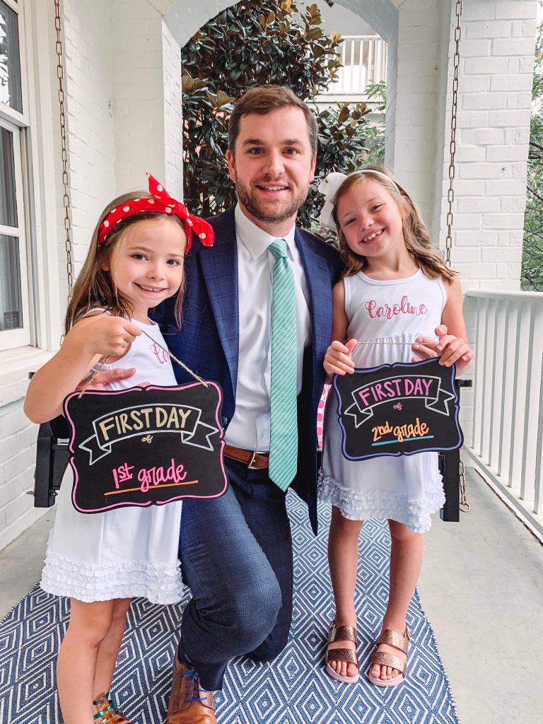 Caroline and Carson Go Back to School... And They're Off! by popular Nashville blog, Hello Happiness: image of two little girls standing outside with their dad and wearing an Elegant Goods Shop dress and holding a Lovely Little Designs chalkboard sign. 