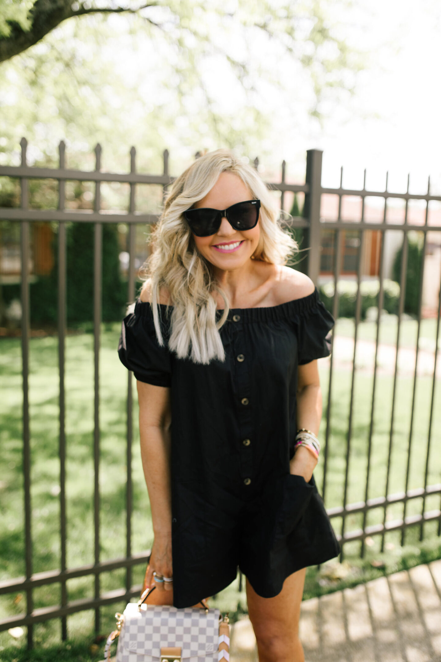 Rompers and Jumpers by popular Nashville fashion blog, Hello Happiness: image of a woman standing in front of a wrought iron fence and wearing a black off the shoulder romper, sunglasses, and holding a grey and white checker handbag. 