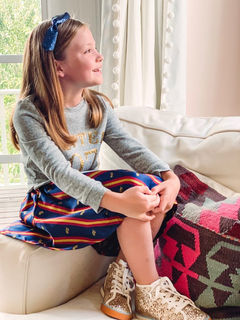 The Magical World of the Mini Boden Harry Potter Collection by popular Nashville fashion blog, Hello Happiness: image of sitting on the arm of a couch and looking out the window  and wearing Mini Boden Sequin Seeker Dress, Mini Boden High Tops, and a blue sequin bow headband.