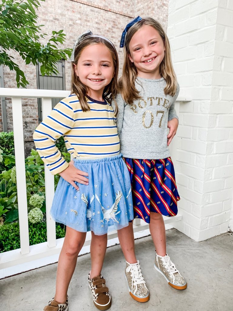 The Magical World of the Mini Boden Harry Potter Collection by popular Nashville fashion blog, Hello Happiness: image of two girls standing next to each other outside with their arms around each other and wearing  Mini Boden Sequin Seeker Dress, Mini Boden High Tops, Mini Boden House Breton - Gryffindor Red/Gold long sleeve shirt, Mini Boden Hippogriff Tulle Skirt, Mini Boden Hippogriff Bomber Jacket, and Fun Low Tops.