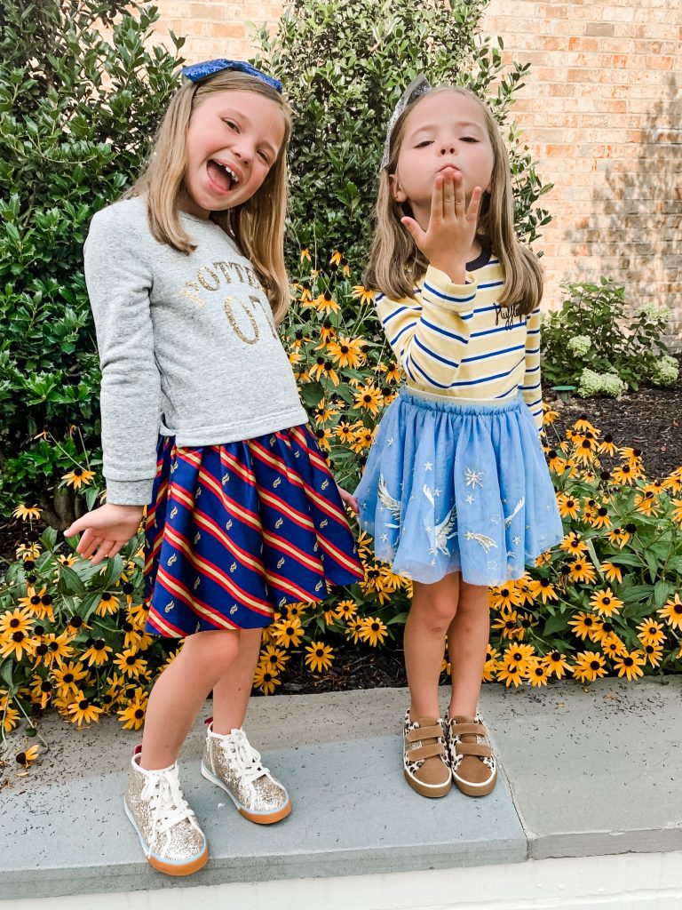 The Magical World of the Mini Boden Harry Potter Collection by popular Nashville fashion blog, Hello Happiness: image of two girls standing next to each other outside with their arms around each other and wearing  Mini Boden Sequin Seeker Dress, Mini Boden High Tops, Mini Boden House Breton - Gryffindor Red/Gold long sleeve shirt, Mini Boden Hippogriff Tulle Skirt, Mini Boden Hippogriff Bomber Jacket, and Fun Low Tops.