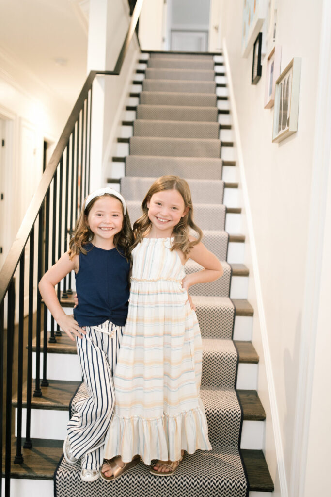 Walmart Back to School by popular Nashville life and style blog, Hello Happiness: image of two young girls standing on a stair case and wearing a Walmart Jessica Simpson Girls Sleeveless Twist Front Top and Paper Bag Pants, Walmart Wonder Nation Shining Star Low-Top Court Sneaker, Walmart Jessica Simpson Girls Halter Neck Striped Maxi Dress, and Walmart bebe Girls Big Kid Two Buckle Strap Cork Footbed Slide Sandals Rose Gold.