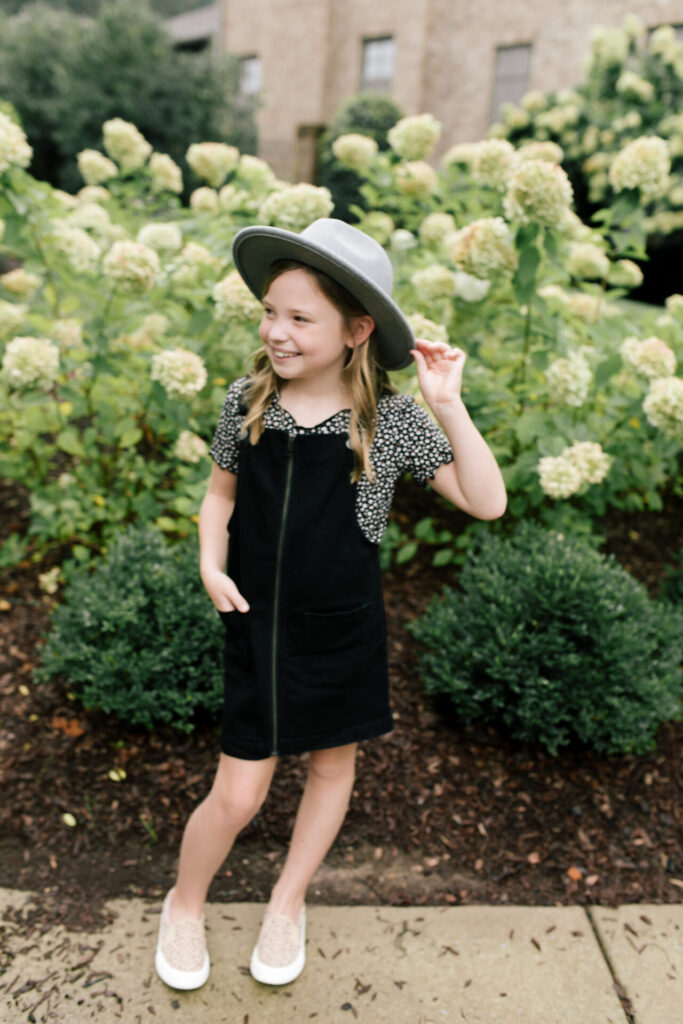 Tween Fashion by popular Nashville fashion blog, Hello Happiness: image of a girl standing outside in front a bush with white flowers and wearing a Target Art Class black pinafore dress, Target thermal floral tee, Target studded sneakers, and Target grey felt floppy studded fedora.