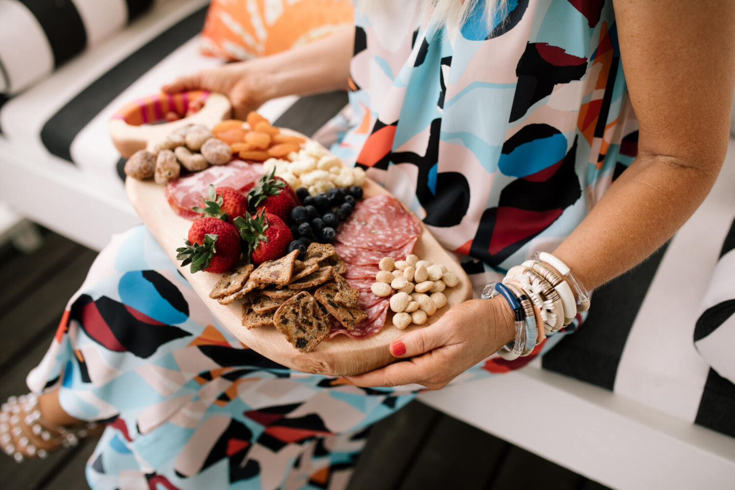 Anthropologie Products by popular Nashville life and style blog, Hello Happiness: image of Natasha Stoneking sitting on a porch swing with black and white stripe cushions and a orange and cream print lumbar pillow and wearing a geometric prints maxi dress and holding a wooden serving board filled with blueberries, yogurt covered raisins, strawberries, salami, dried apricots and nuts. 