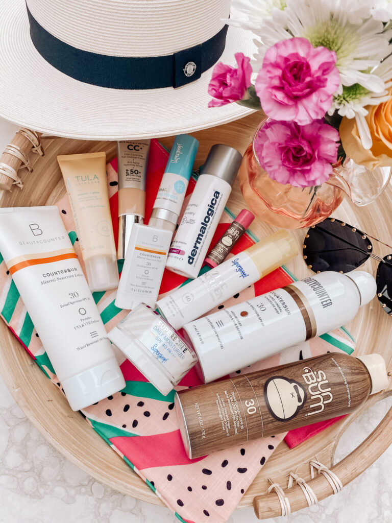 Sun Protection Products by popular Nashville beauty blog, Hello Happiness: image of a white woven beach hat, a vase of pink and white flowers, studded aviator sunglasses, watermelon wet bag, bamboo serving tray, and sun-bum spray on sunscreen, Tula sunscreen, dermalogica sunscreen, Supergoop sunscreen and moisturizer, itcosmetics cc cream, and Beautycounter sunscreen. 