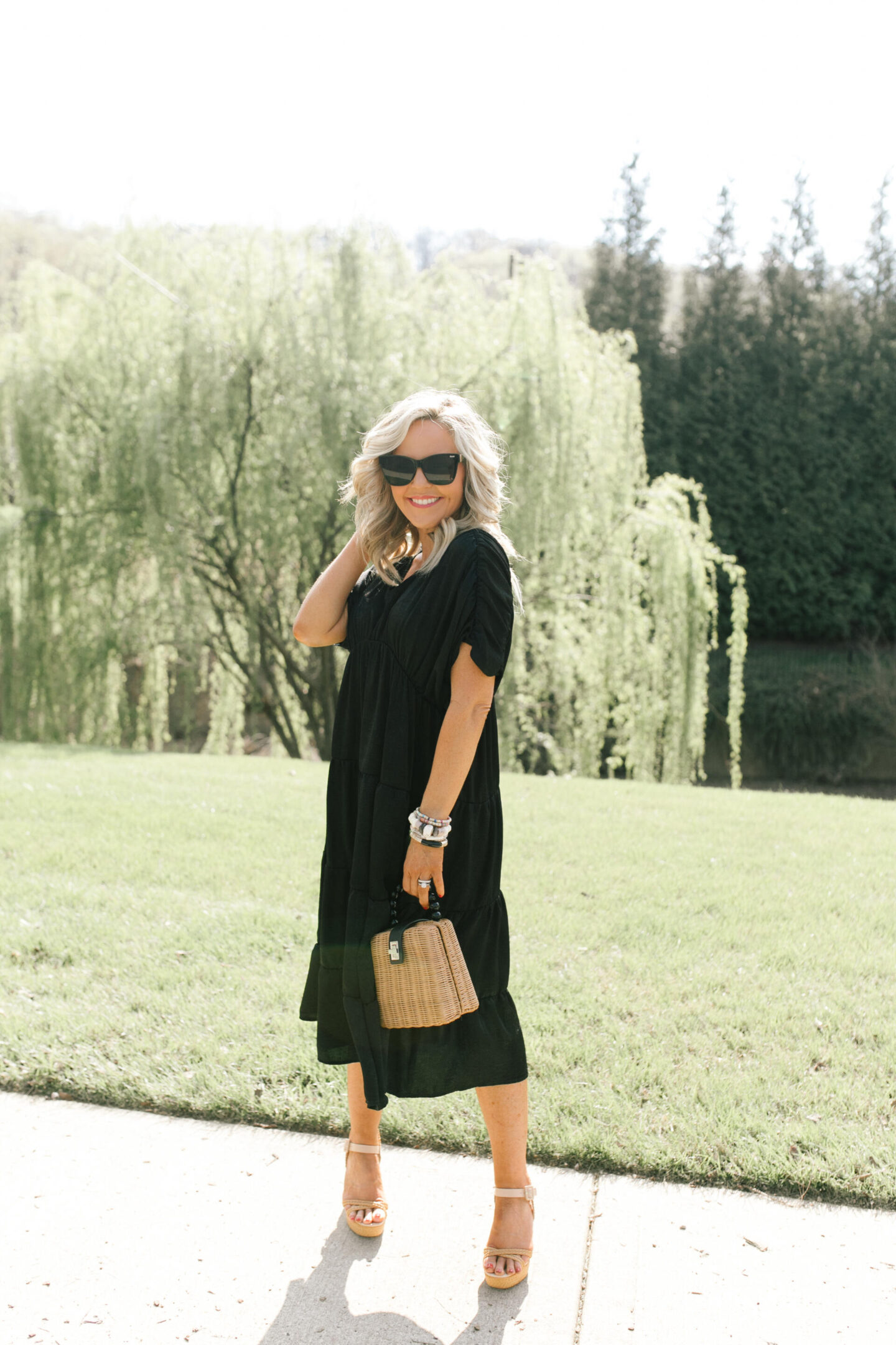 Summer Dresses by popular Nashville of Natasha Stoneking wearing a shirred tiered midi dress, after hours sunglasses, platform wedge sandals, and holding a woven bucket bag. 