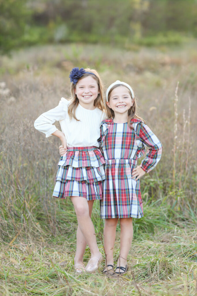 Gift Ideas for Girls by poplar Nashville life and style blog, Hello Happiness: image of two young girls standing next to each other in a field and wearing a white blouse, gold ballet flats, blue sandals, blue flower headband, white knot head band, plaid tier skirt, and plaid dress. 