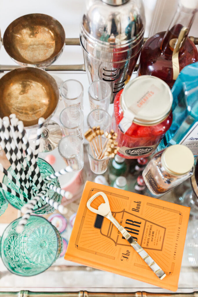 Bar Cart Essentials by popular Nashville lifestyle blog, Hello Happiness: image of a Ballard Designs Jill Bar Cart, Pottery Barn Harrison Bottle Coaster, Amazon The Essential Bar Book: An A-to-Z Guide to Spirits, Cocktails, and Wine, with 115 Recipes for the World's Great Drinks, gold drink stirs, bold bottle opener, black and white straws, and clear shot glasses. 